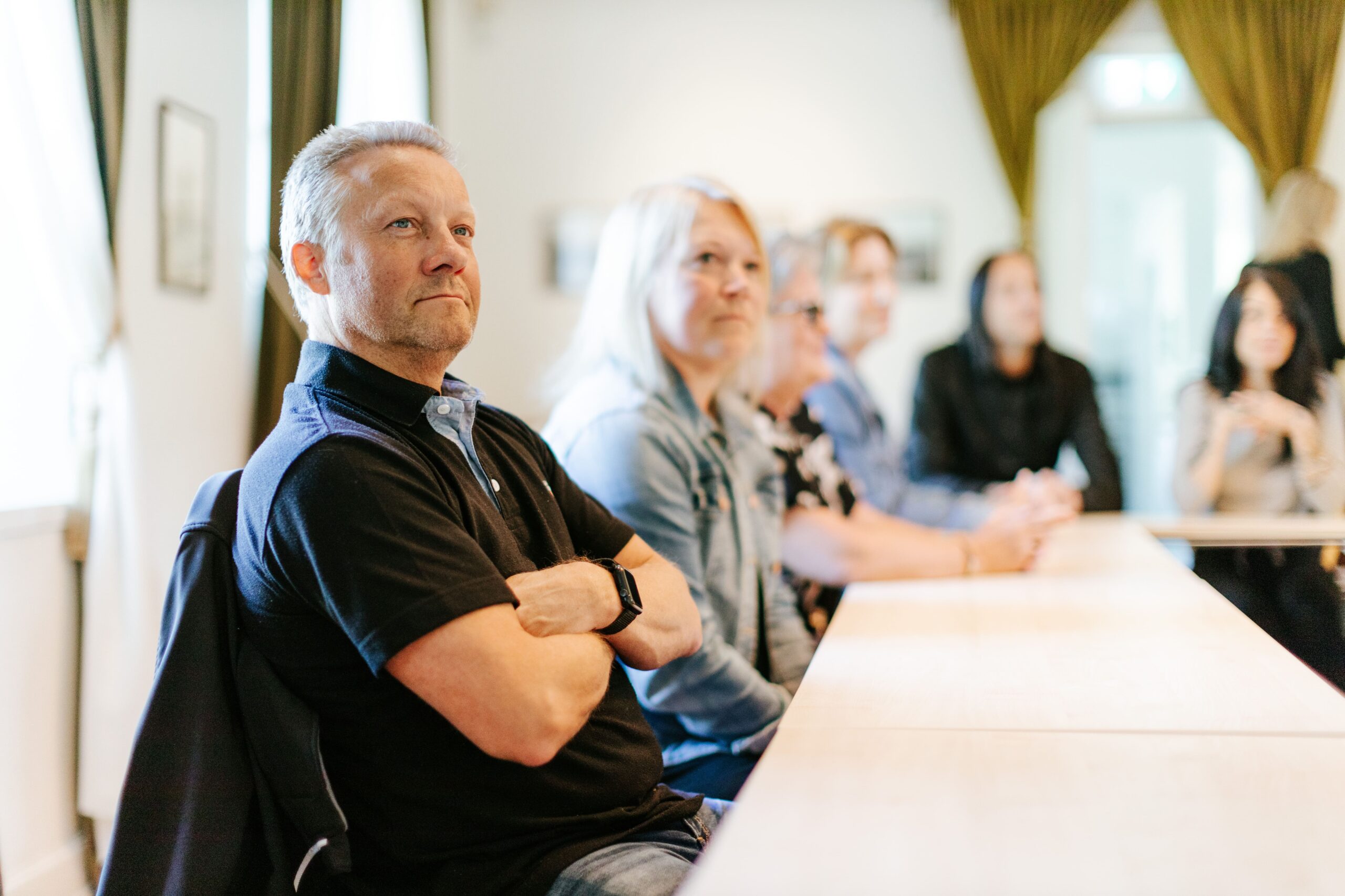 Konferensdeltagare i en ljus lokal. Fokus på en man i förgrunden, med uppmärksamhet på diskussionen framme vid bordet.
