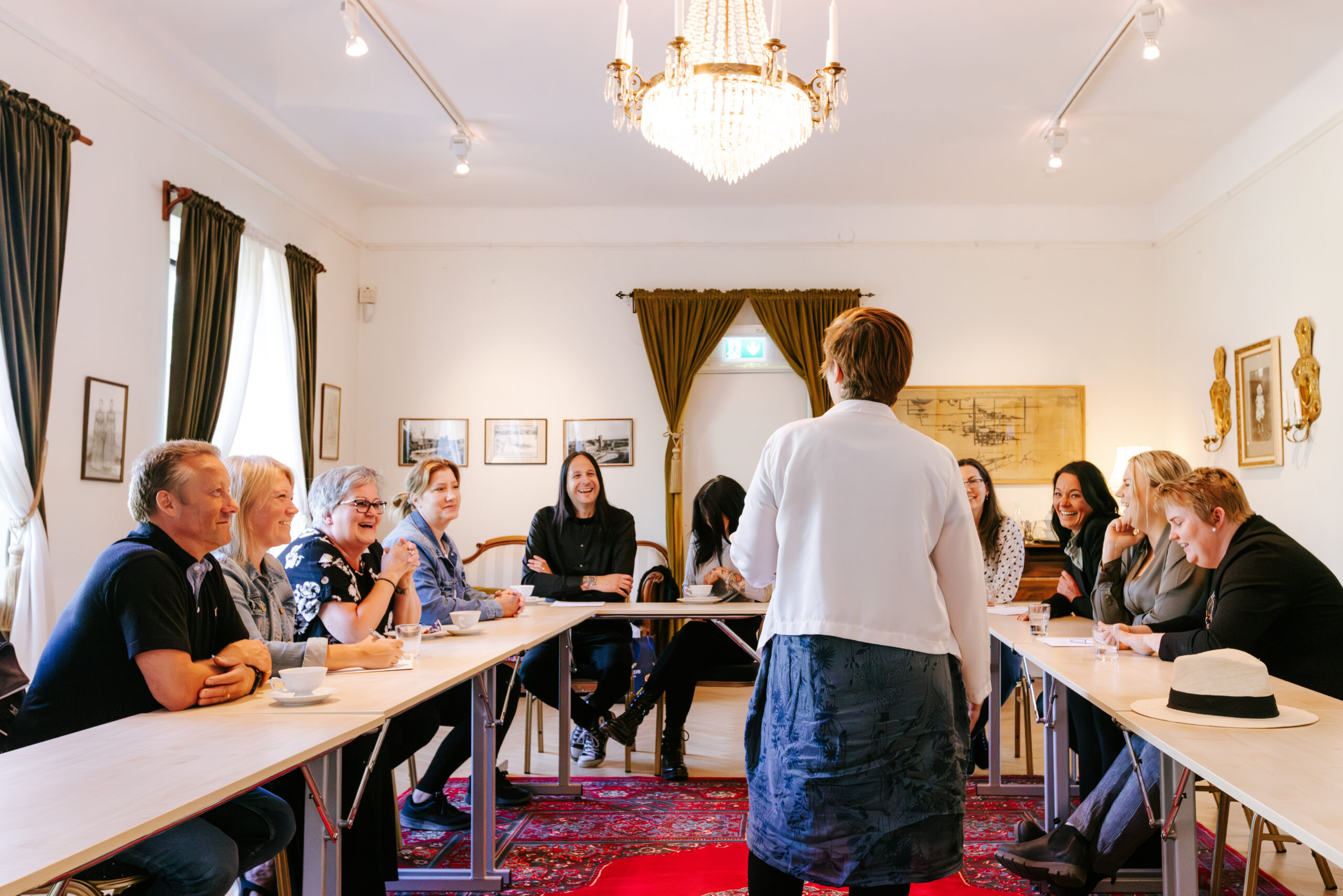 Möte i en ljus konferenslokal med deltagare som diskuterar, föreläsare framför och en ly chandelier i taket.