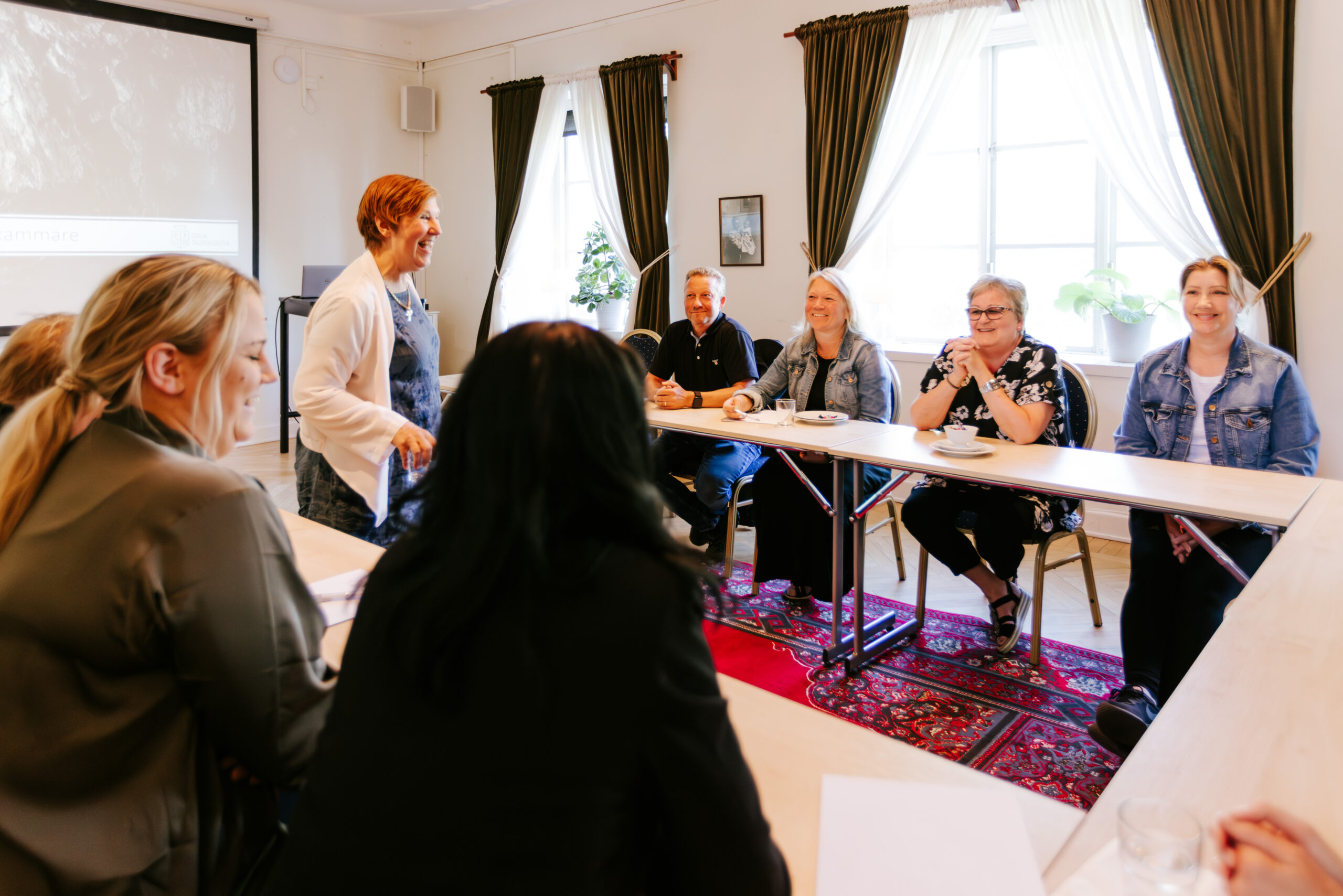 Konferensdeltagare sitter vid ett bord, ler och lyssnar på en talare i en ljus och inbjudande miljö.