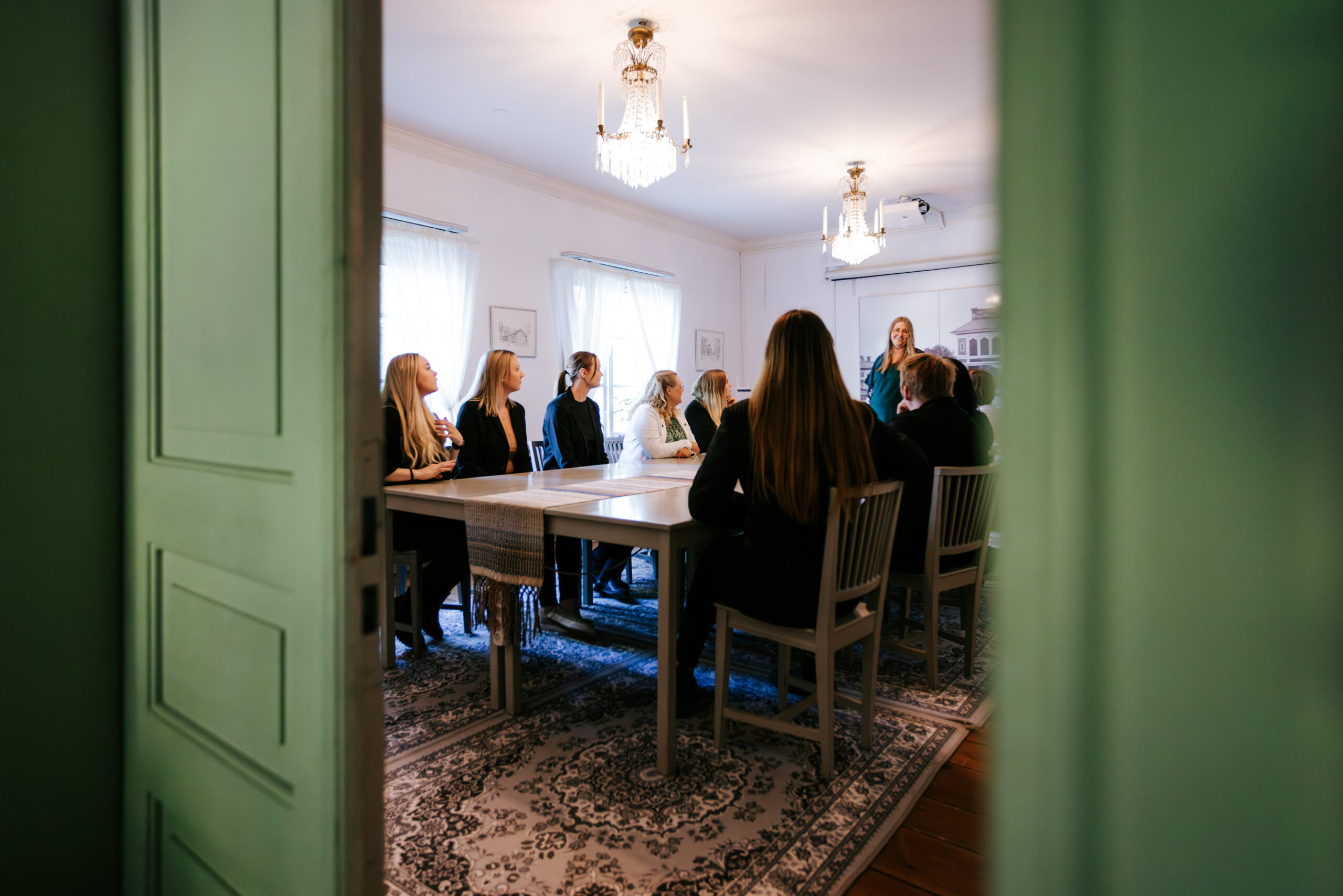 Möte i en elegant sal med kvinnor som lyssnar på en talare vid ett långbord. Ljus, neutral färgpalett och elegant atmosfär.