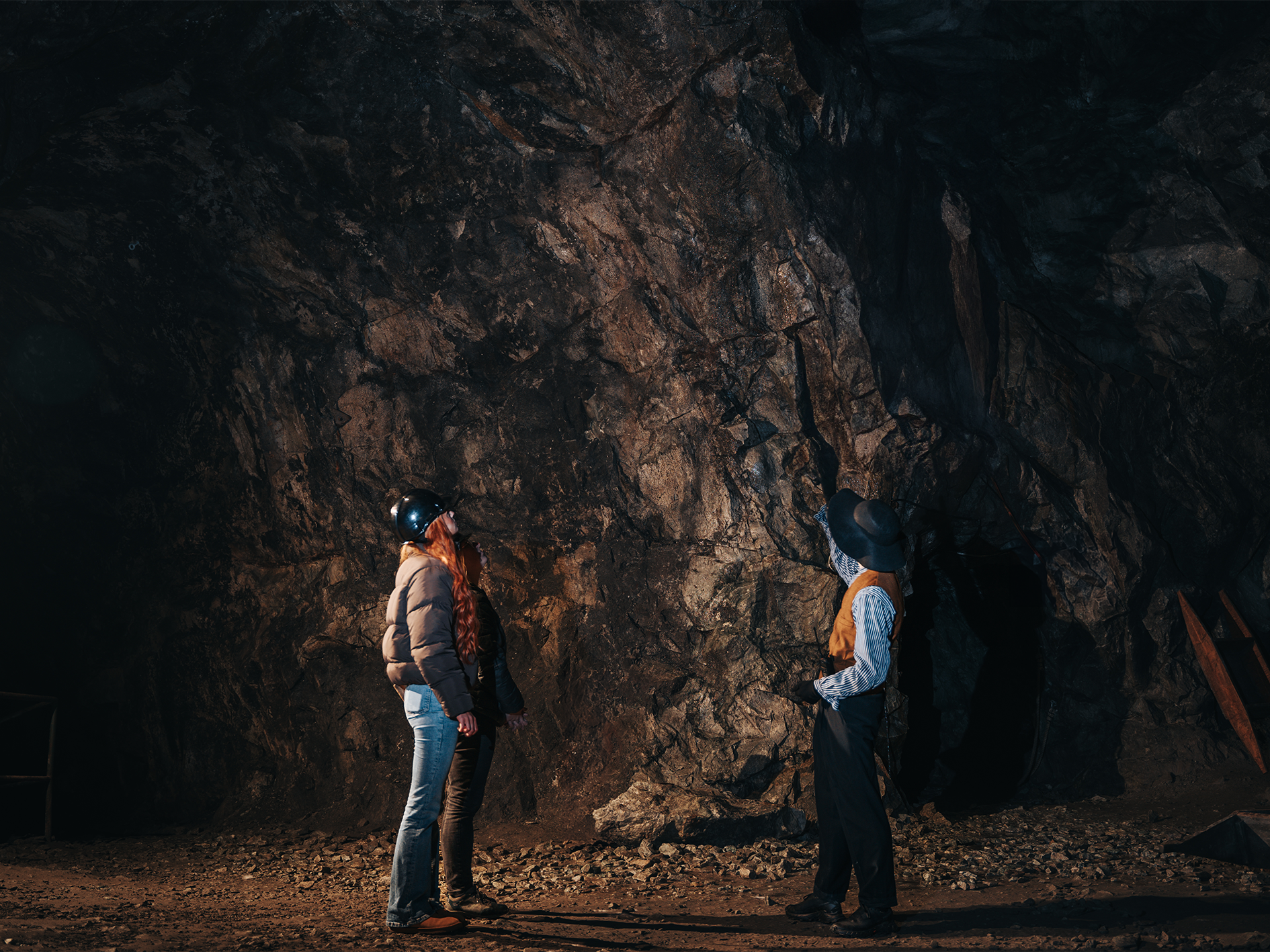 Två personer i hjälmar undersöker en bergvägg i Sala Silvergruva, omgiven av mörka stenar och gruvutrustning.