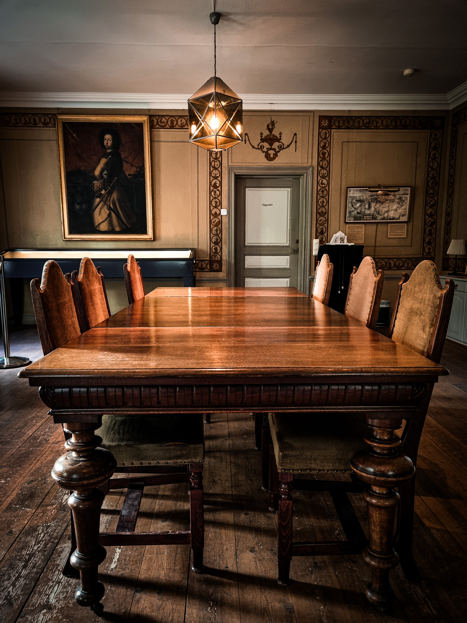 Alt-text: Ljust rum med träbord och stolar, en elegant lampa hänger ovan, samt en målning på väggen. Passar utställningar i Sala Silvergruva.