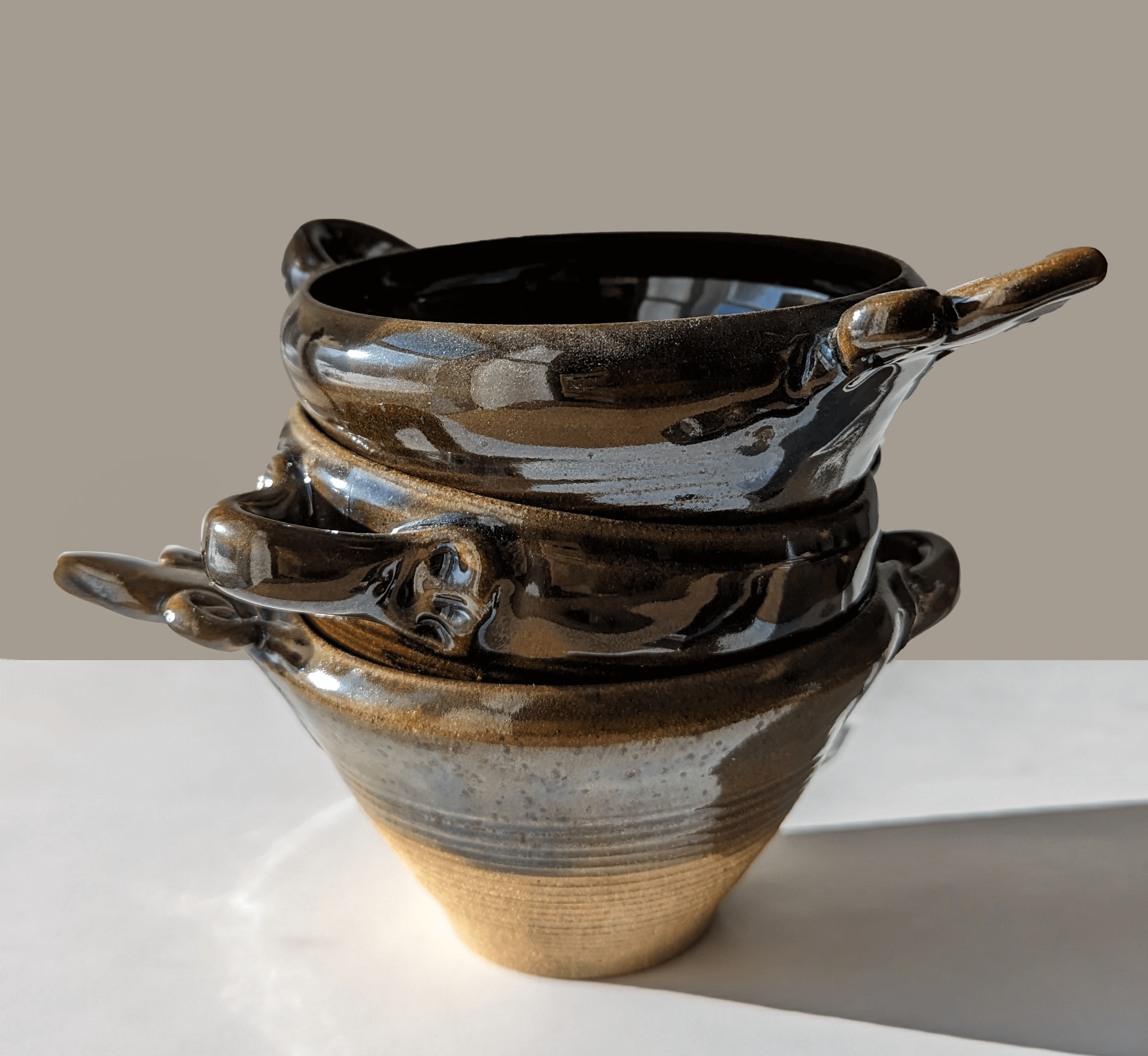 Tre staplade gruvbyskålar med glänsande ytor, svart och bronsfärg med detaljerade handtag, mot en neutral bakgrund.