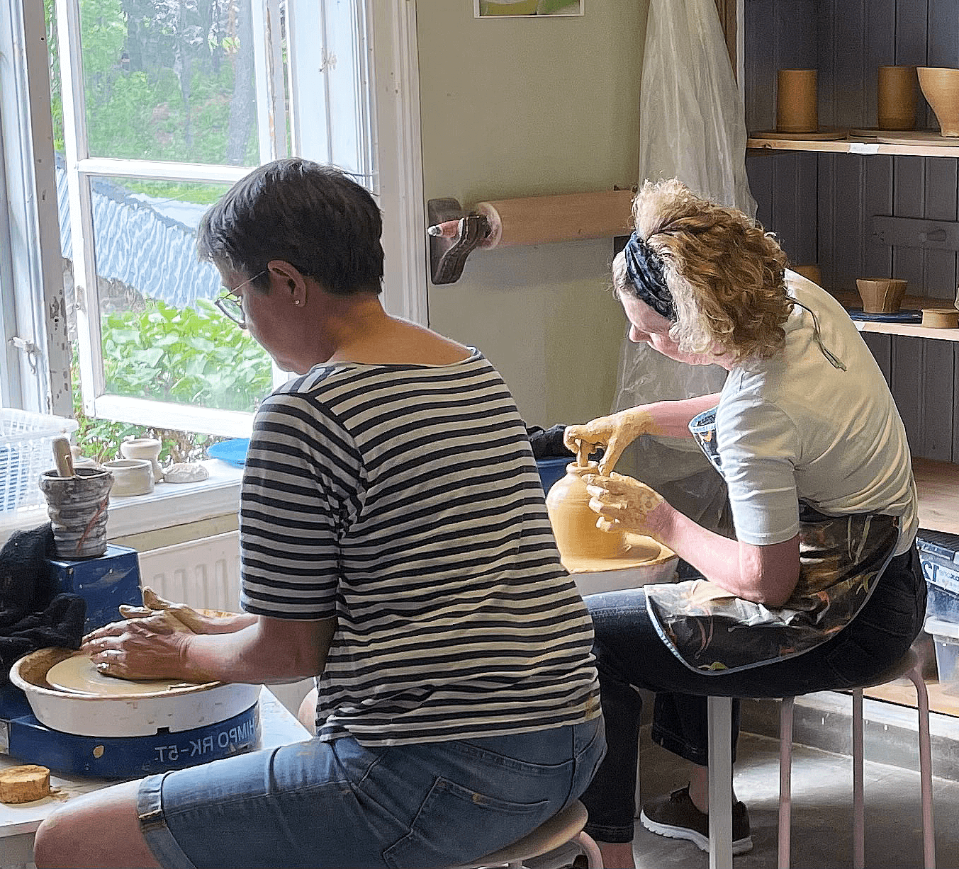 Två kvinnor drejar i ett ljust rum med fönster, omgivna av keramiska föremål och verktyg. Kreativ atmosfär och tradition.