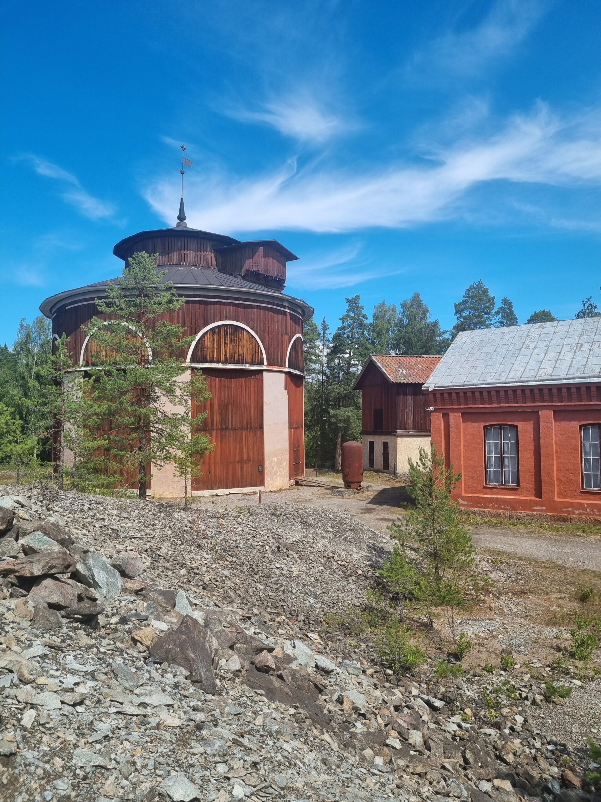 Runda byggnaden i Sala Silvergruva med träfasad och omgivande natur under en klar blå himmel.