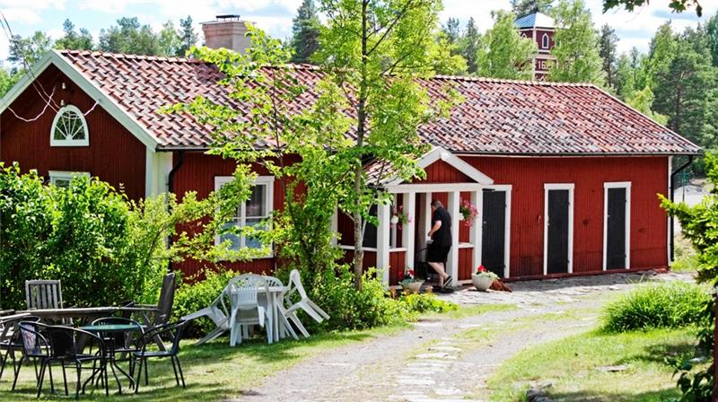 Röd stuga med takpannor, omgiven av grönska och träd, inbjudande entré och utemöbler för avkoppling.